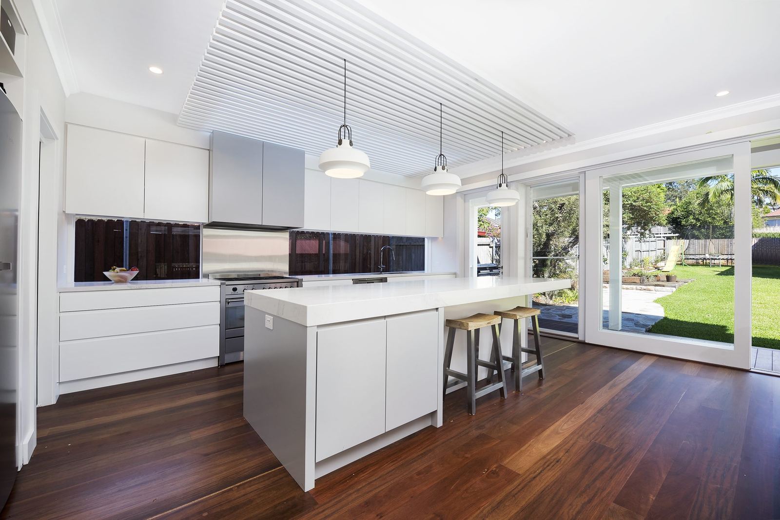Seaforth — white kitchen island with pendant lights and sliding doors to garden, built by HP Constructions with Playoust Churcher